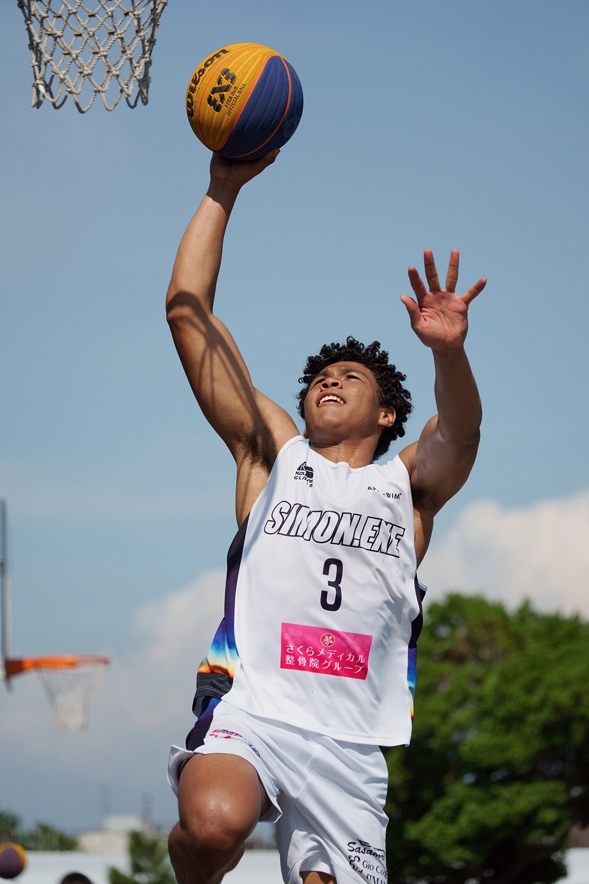 Jugador de baloncesto en pleno vuelo durante un partido al aire libre, capturado desde un ángulo bajo contra un cielo azul: un ejemplo enérgico de fotografía deportiva dinámica que enfatiza el movimiento y la intensidad.
