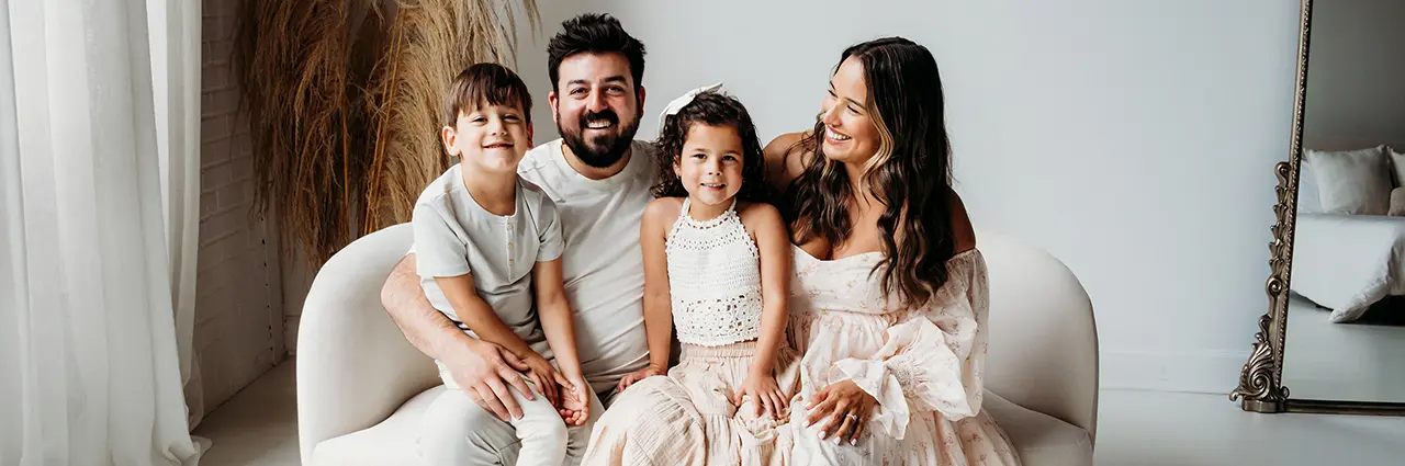 Familia sentada junta en un sofá blanco, sonriendo durante una sesión de fotos - ejemplo de objetivo de focal fija frente a objetivo zoom para fotografía familiar.