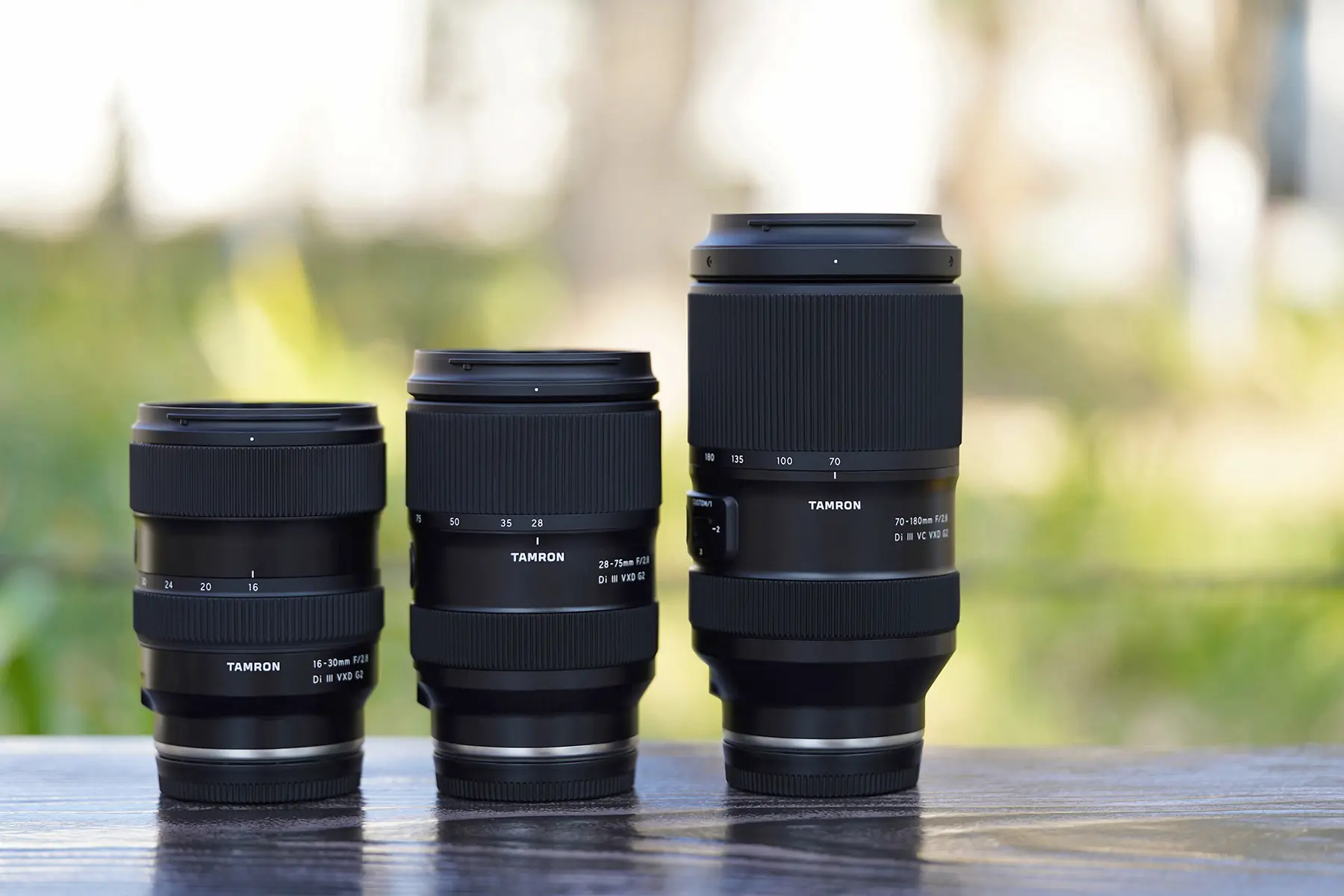 Three Tamron Di III VXD G2 lenses—a 16-30mm, 28-75mm, and 70-180mm—lined up side by side on a wooden surface with a softly blurred outdoor background, highlighting their compact design and focal range coverage.