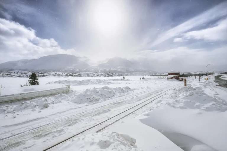A bright winter sun hovers over a snowy mountain landscape and railroad tracks leading toward a small red building, with snowflakes drifting in the crisp air and dramatic clouds stretching across the sky.
