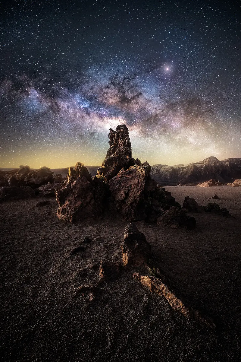 Formação rochosa imponente iluminada sob a Via Láctea, fotografada com uma lente Tamron f/2.8 rápida - um exemplo do brilho e da clareza da melhor lente para astrofotografia.