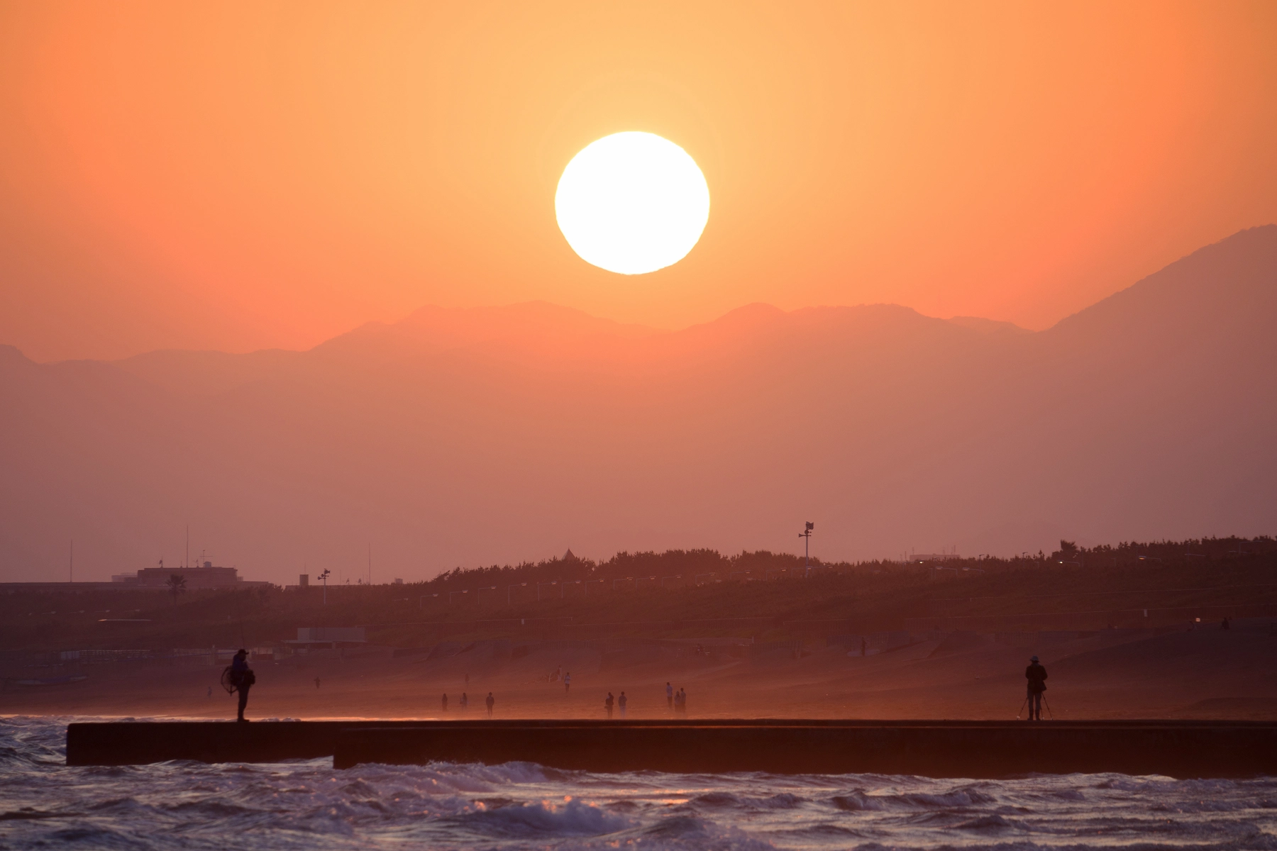 Puesta de sol sobre un paisaje costero con siluetas de fotógrafos, montañas y olas del océano, captada con uno de los mejores objetivos Nikon Z para fotografía de paisajes.
