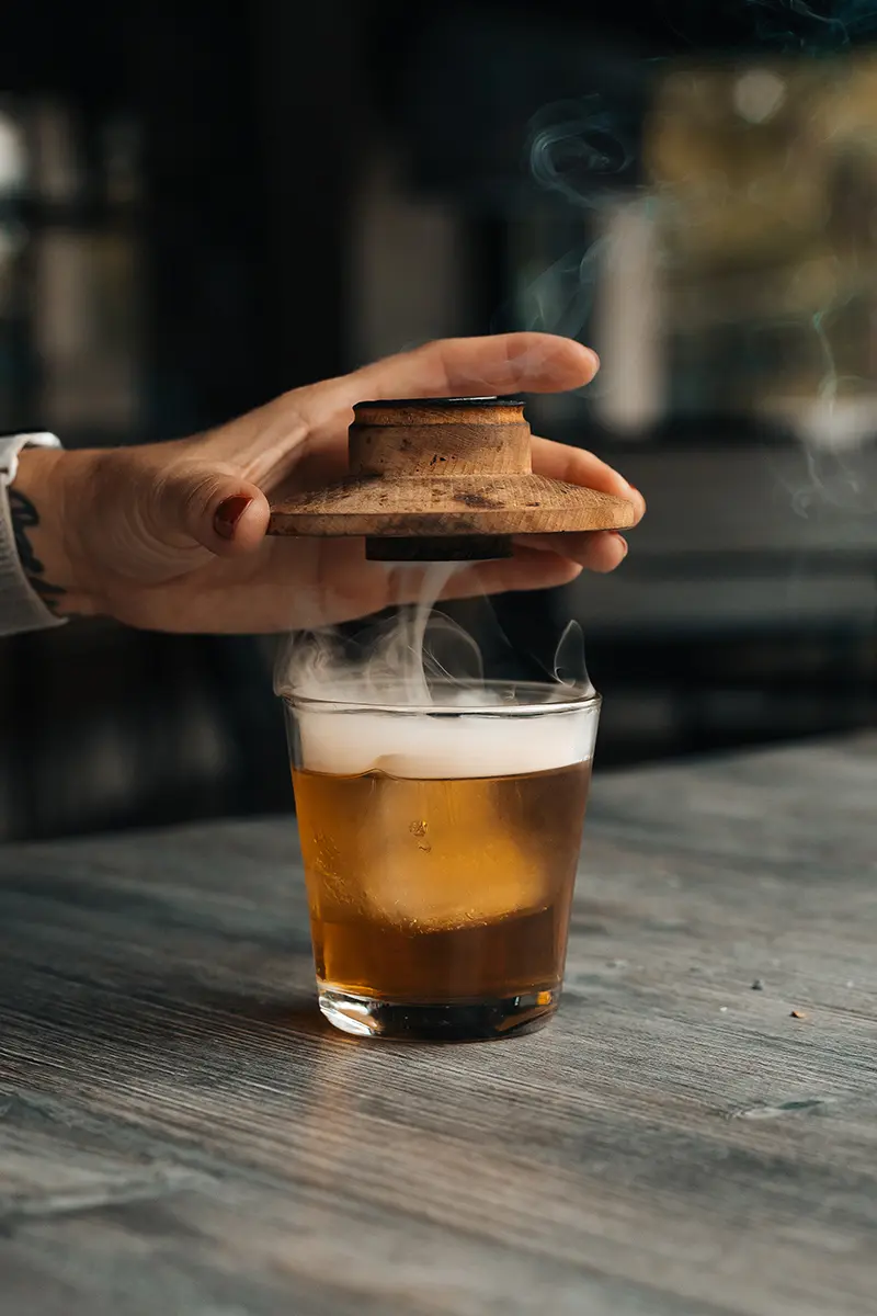 Levantando a mano una tapa de madera de una copa de cóctel ahumada con vapor ascendente, capturado para la fotografía de alimentos y bebidas de otoño.