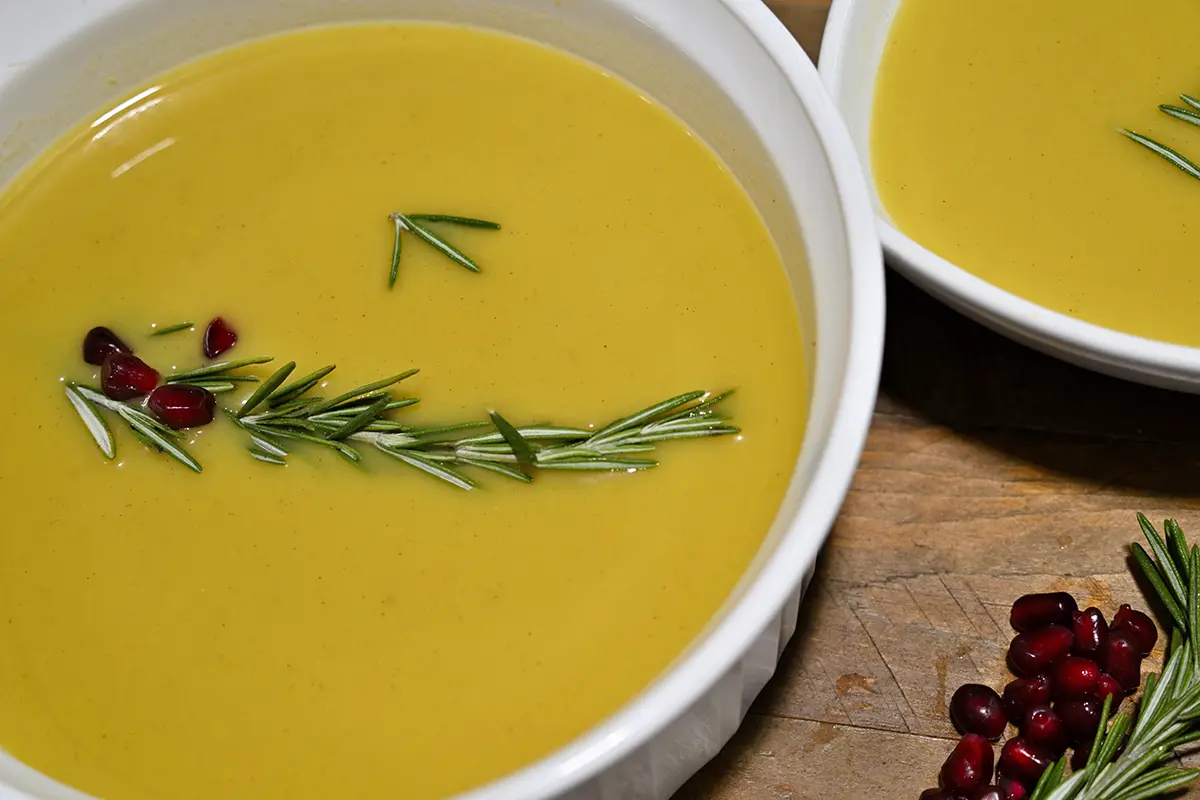 Tazón de sopa de calabaza aderezado con romero y granos de granada, en una mesa rústica.