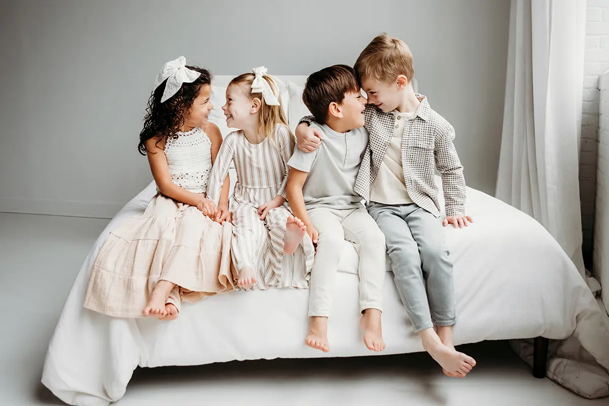Quatro crianças pequenas sentadas juntas em uma cama, sorrindo e interagindo durante uma sessão de fotos - exemplo de lente prime versus zoom para fotografia de família.