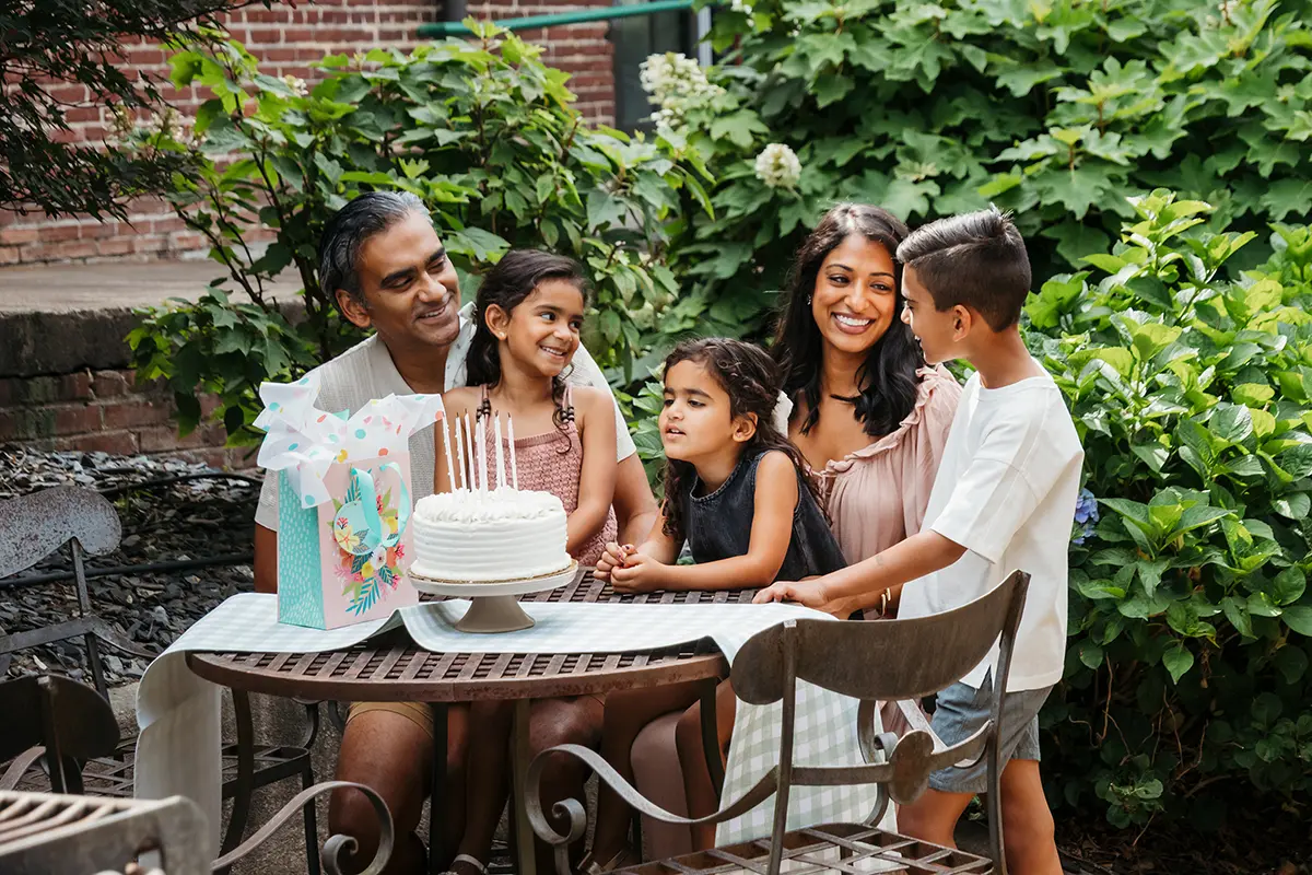 Família comemorando o aniversário de uma criança ao ar livre, reunida em torno de um bolo com velas acesas e sorrindo juntos.