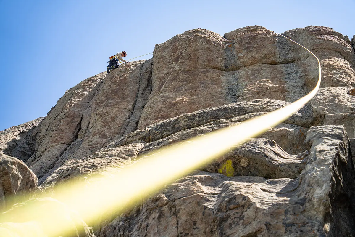 Grimpeur escaladant une falaise rocheuse avec une corde jaune menant vers l'appareil photo sous un ciel bleu clair.