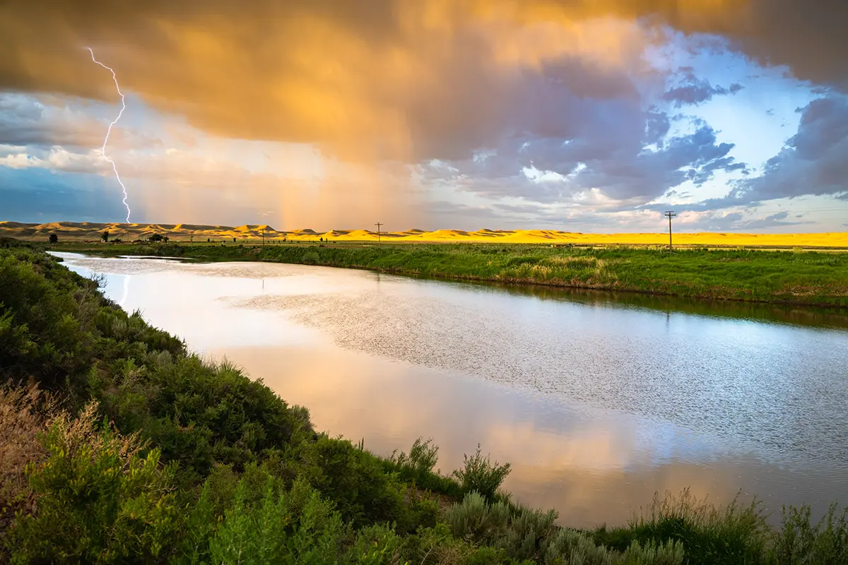 La foudre frappe les champs dorés et la rivière au coucher du soleil, capturant des moments dramatiques pour une mission de conseils en photographie de voyage.