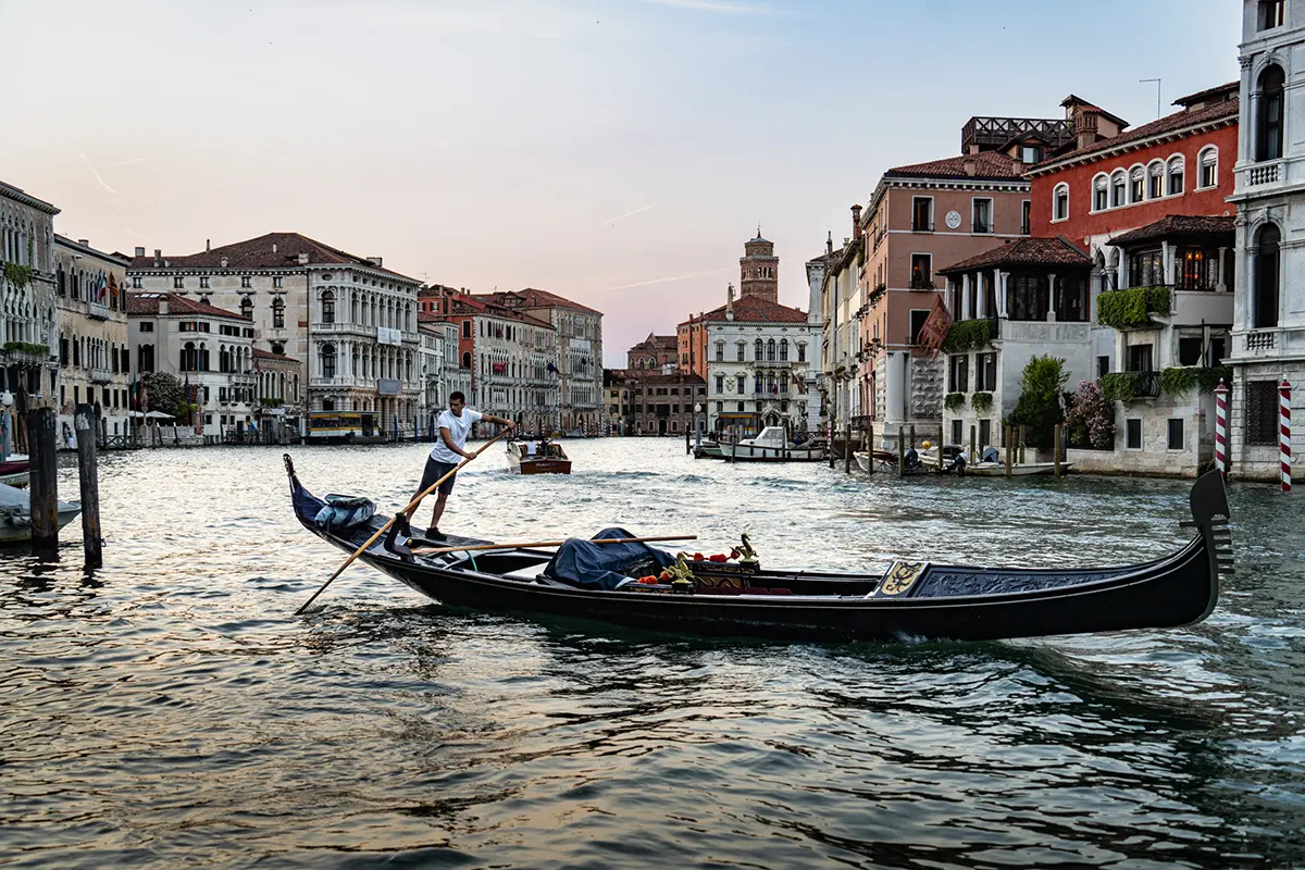 Un gondolier fait avancer une gondole traditionnelle sur le Grand Canal de Venise au coucher du soleil, entouré de bâtiments historiques.
