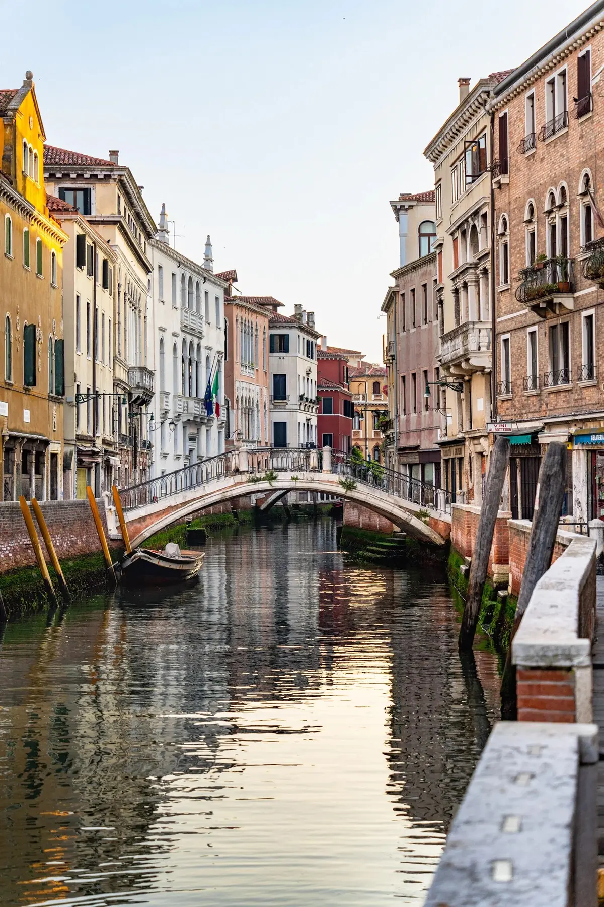 Une passerelle en pierre enjambe un canal vénitien tranquille au coucher du soleil, avec les reflets des bâtiments historiques dans l'eau.