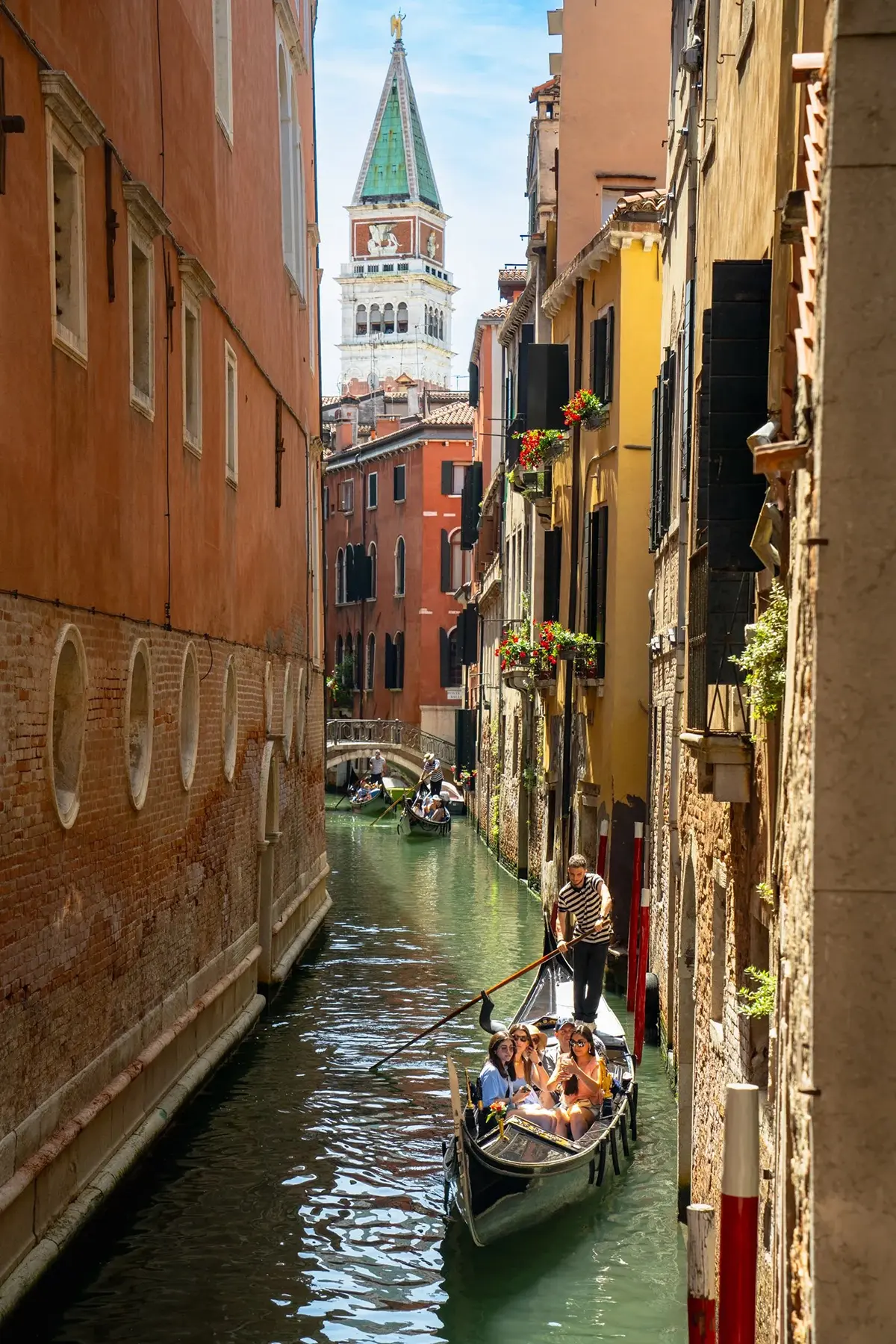 Un gondolier conduit des touristes à travers un étroit canal vénitien, avec la tour Campanile di San Marco en arrière-plan.
