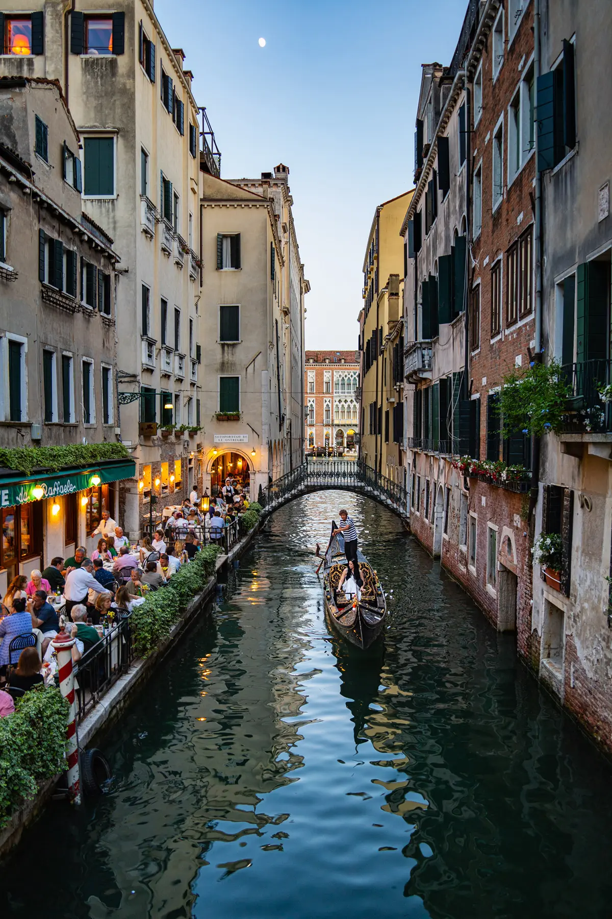 Un gondolier passe devant un restaurant animé au bord d'un canal à Venise au crépuscule, les convives profitant de l'atmosphère de la soirée.