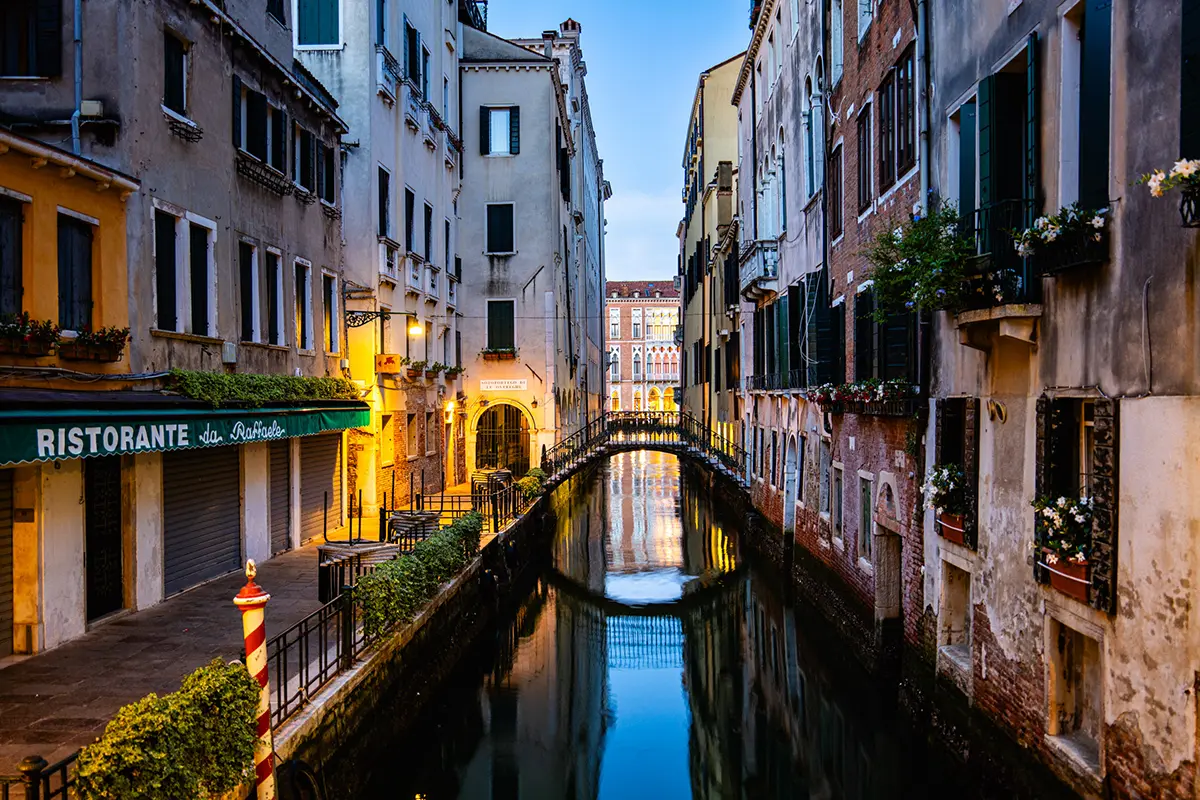 Vue nocturne d'un canal vénitien tranquille avec des lumières chaudes se reflétant sur l'eau et une petite passerelle au loin.