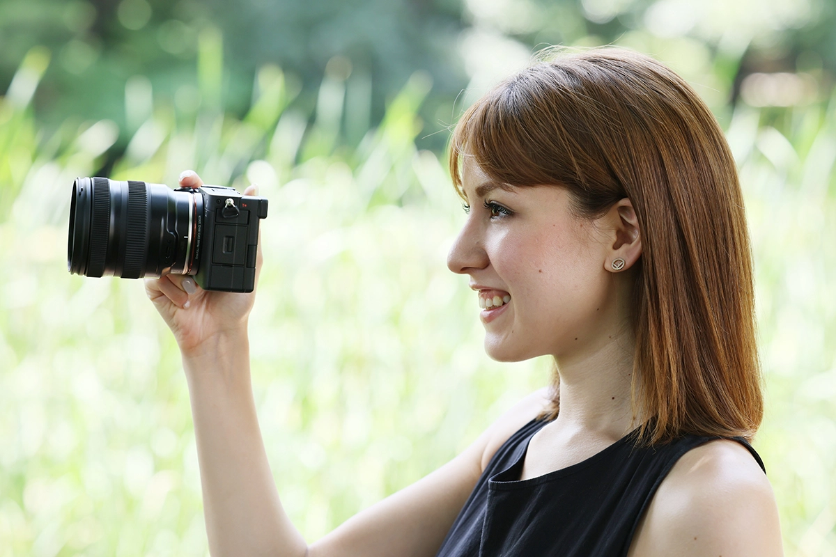 Mujer sonriente filmando con una cámara compacta sin espejo y objetivo zoom en exteriores, sosteniéndola en posición selfie o vlogging con un fondo verde difuminado.