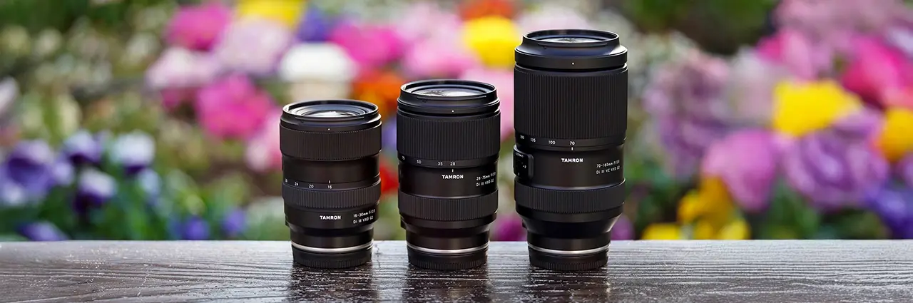 Tamron trinity lenses lineup — 16-30mm, 28-75mm, and 70-180mm F2.8 G2 zoom lenses for Sony and Nikon mirrorless cameras, displayed side by side outdoors with colorful bokeh background.