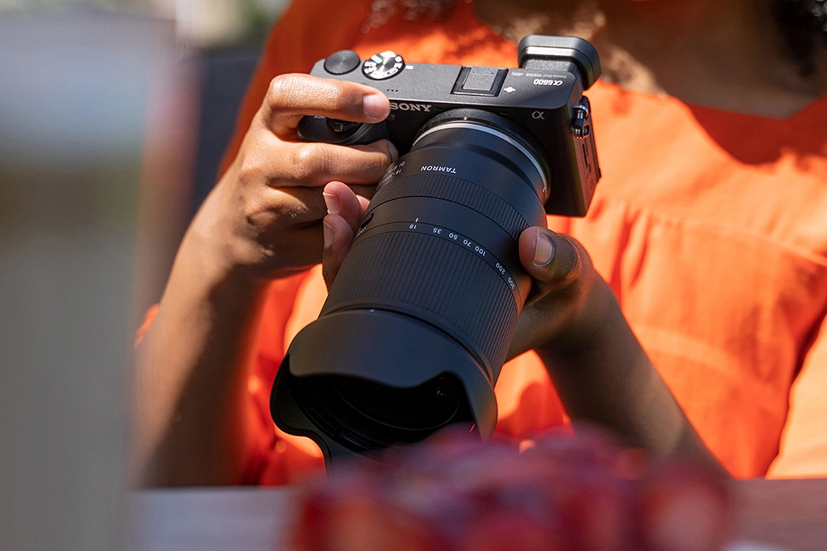 Mãos segurando uma câmera Sony com lente Tamron 18-300 mm durante uma sessão de fotografia de família ao ar livre.