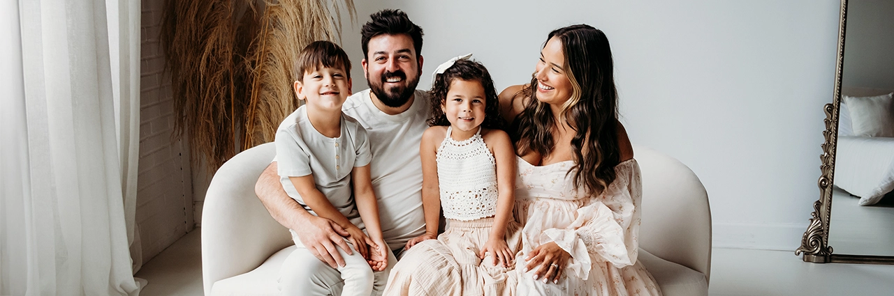 Família sorridente sentada em um sofá branco durante uma sessão de fotografia de família em ambiente interno com luz natural de janela.
