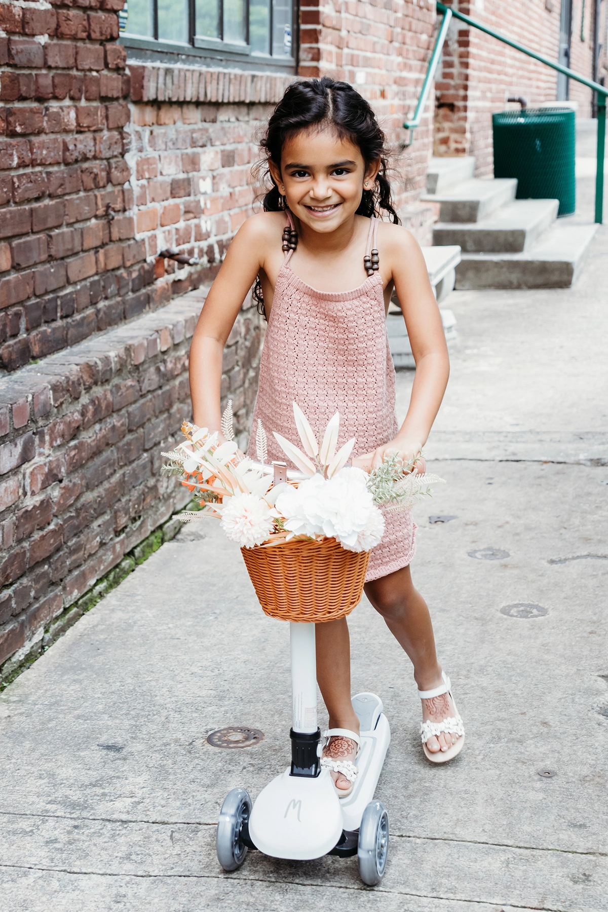 Menina sorridente andando de scooter com flores na cesta durante uma sessão de fotografia de família ao ar livre.