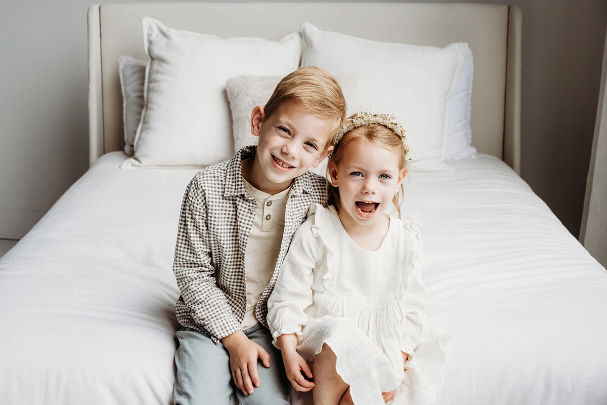 Irmão e irmã sentados em uma cama perto da janela, sorrindo durante uma sessão de fotografia de família em ambiente interno.