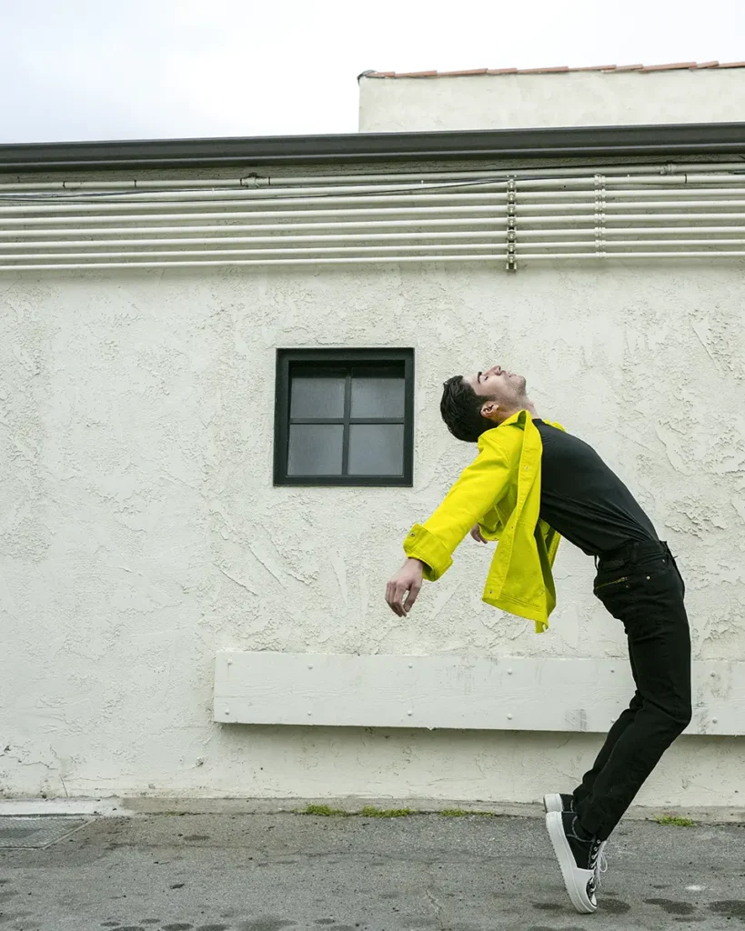 A young man with yellow and black on bending backwards set against a white wall outside.