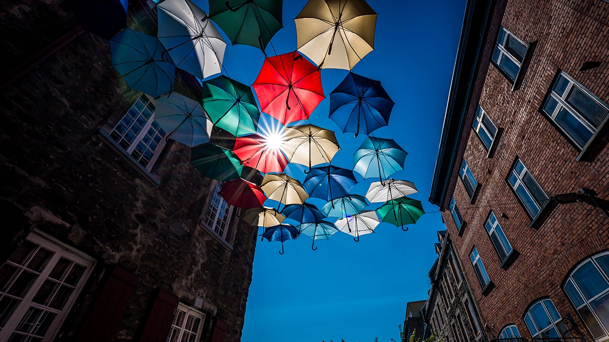 Una vibrante escena callejera con paraguas de colores suspendidos en lo alto, formando un dosel contra un cielo azul despejado, con los rayos del sol visibles a través de ellos y enmarcados por viejos edificios de ladrillo a ambos lados, con múltiples ventanas. 