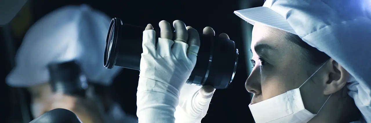 Tamron technician inspecting a camera lens during quality control in a cleanroom environment.
