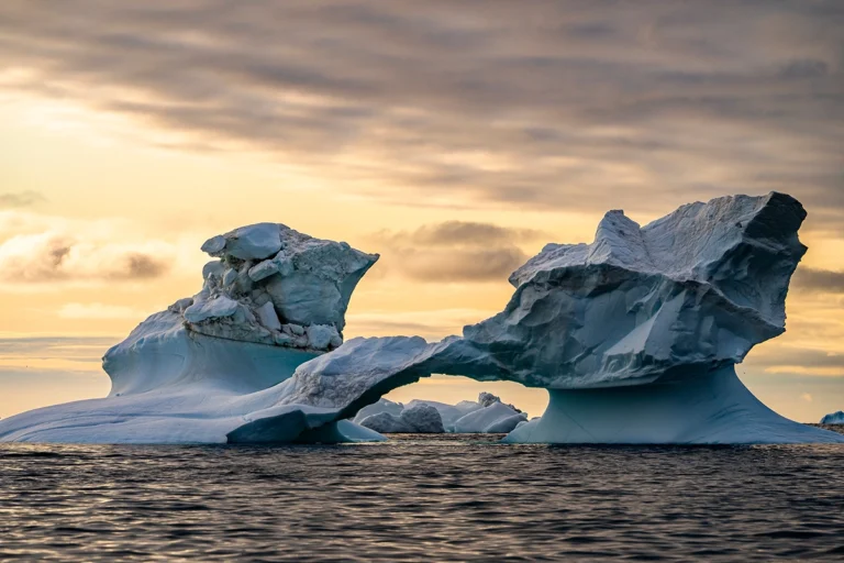 Dois icebergs grandes e esculpidos flutuando no oceano sob um céu dramático ao pôr do sol.