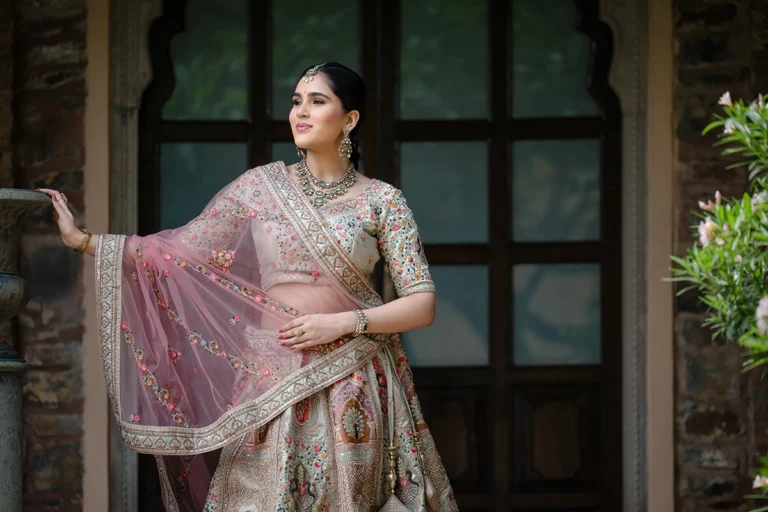 Uma mulher vestida com uma lehenga indiana tradicional, em pé ao ar livre, com um edifício com uma porta em arco e portas de madeira atrás dela.