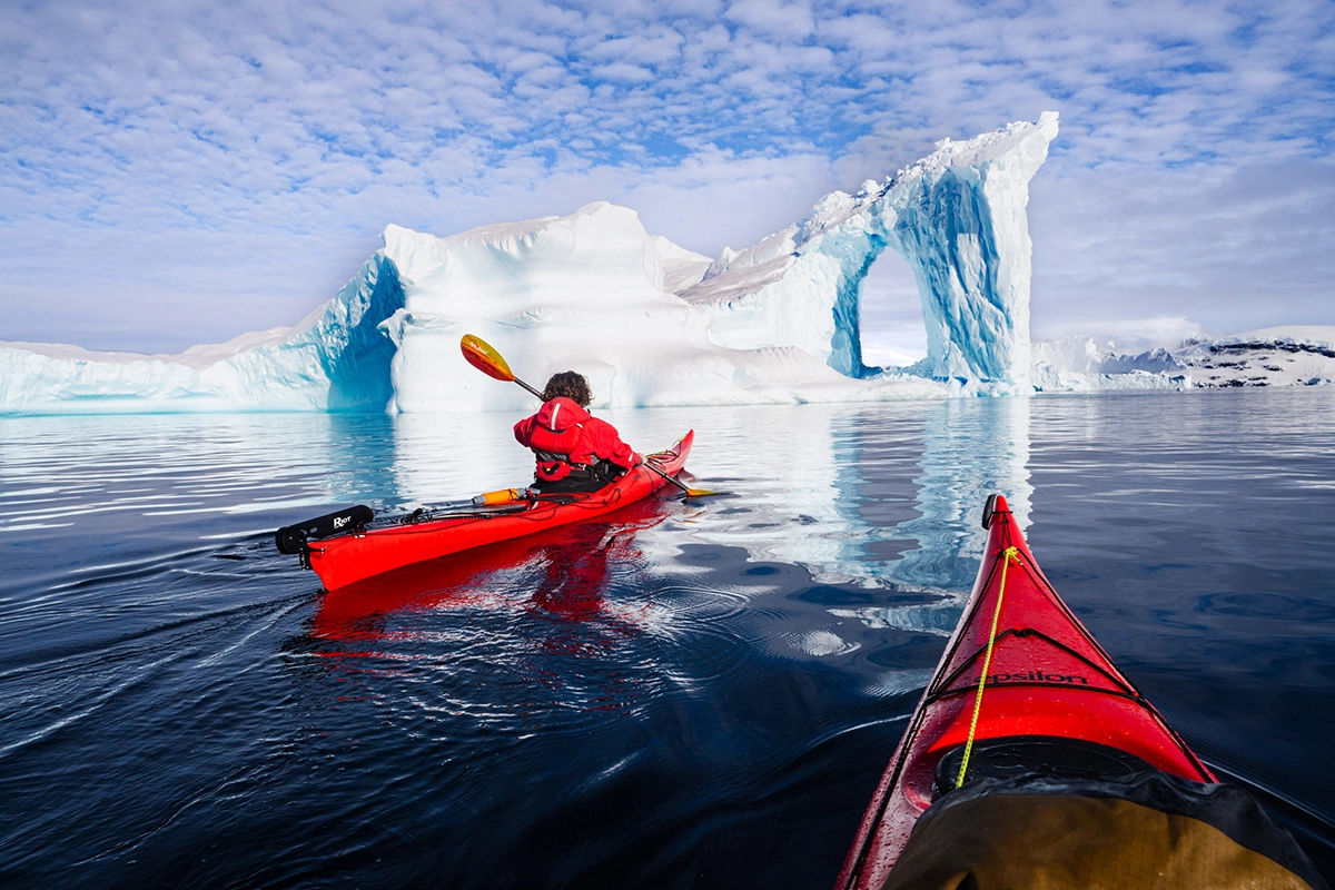 Kayaker in a red kayak paddling toward a massive arch-shaped iceberg during an Antarctica cruise photography expedition.