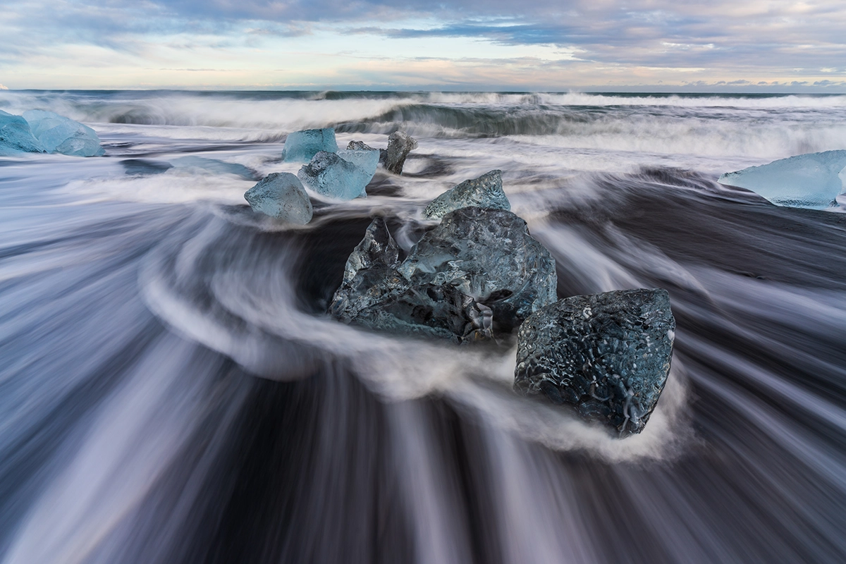 Ice chunks on a black sand beach with waves swirling around them, demonstrating dramatic composition and motion blur techniques for photographers entering photo contests.