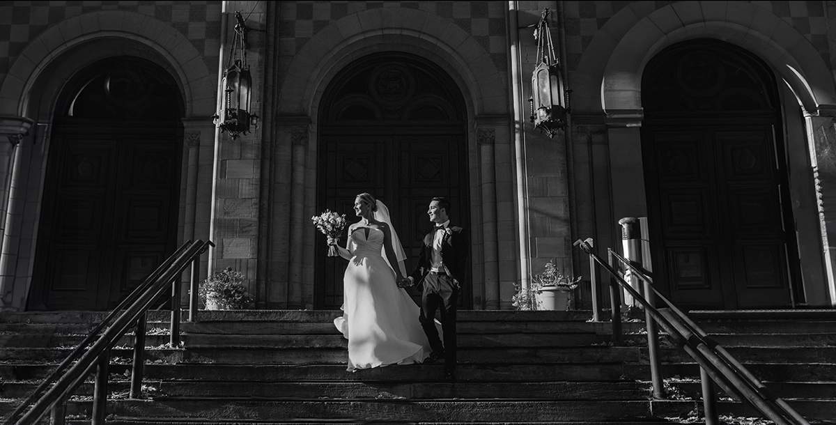 Black and white photo of a bride and groom walking down church steps in elegant lighting, emphasizing contrast and storytelling.