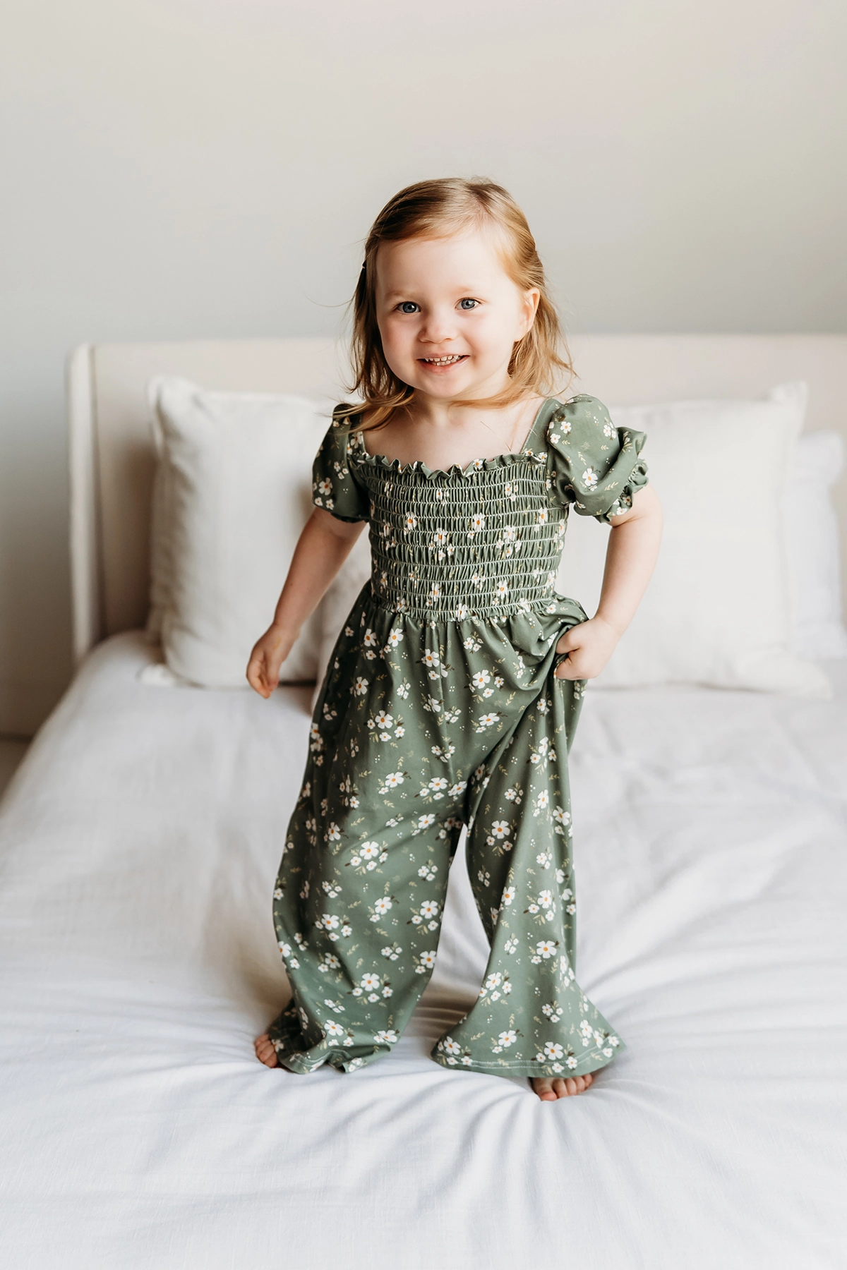 Smiling young girl in a green floral outfit jumping on a bed in natural light, capturing joyful emotion and candid portrait techniques.