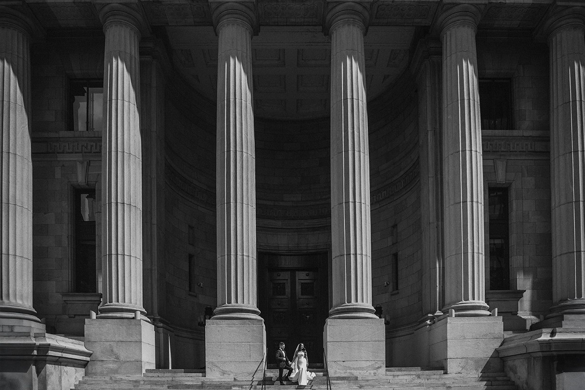 A wedding couple sits on the steps of a grand building with tall stone pillars.