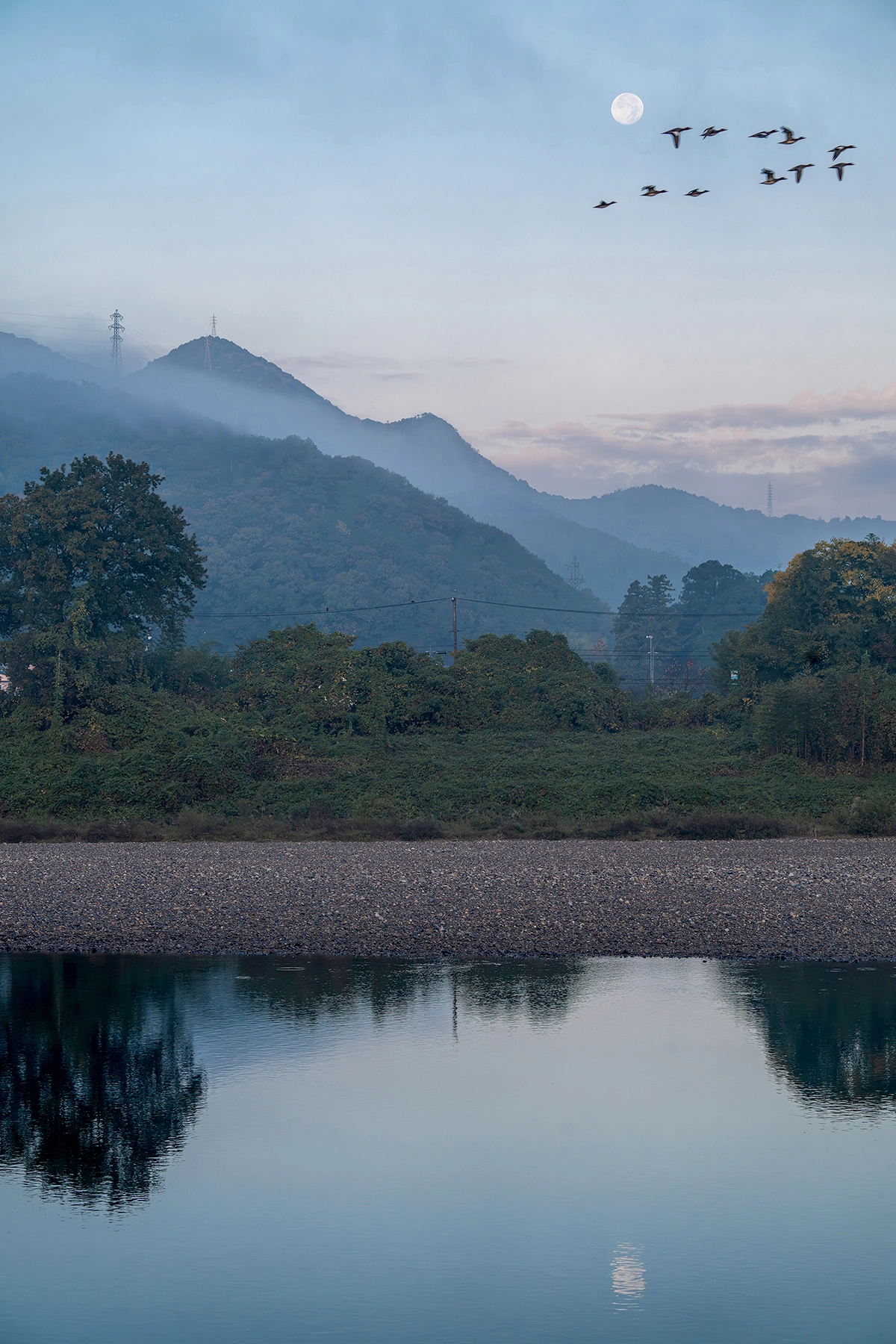 Full moon in daytime sky above misty mountains with birds flying across.