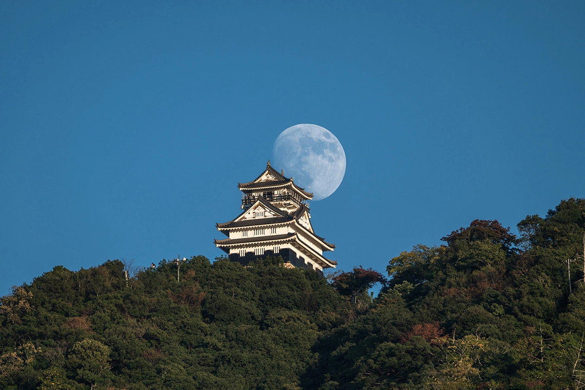 Full moon in daytime sky positioned above a Japanese castle on a hill.