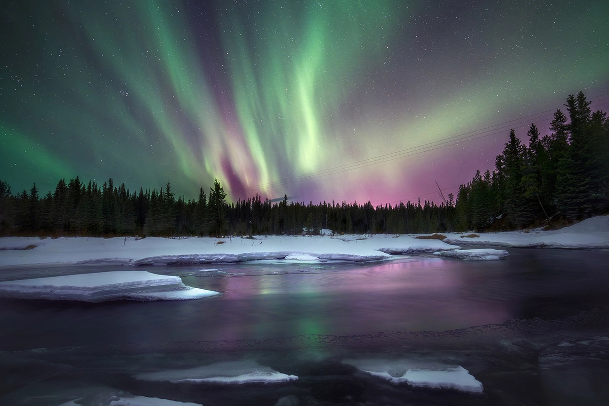 Una aurora boreal púrpura y verde se refleja en una tranquila masa de agua rodeada de nieve.