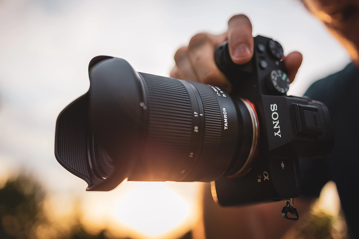 Primer plano de un fotógrafo que sostiene una cámara Sony equipada con un objetivo Tamron, captado con luz solar cálida y destellos del objetivo, lo que ilustra la calidad y la sensación de una mejora profesional del objetivo.