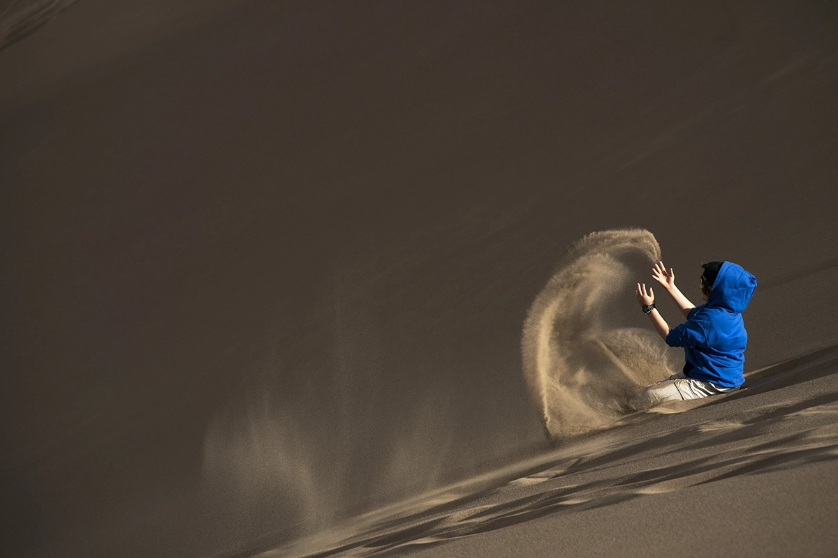 Personne vêtue d'un sweat à capuche bleu vif jouant avec le sable sur une dune, créant un fort contraste de couleur et de texture pour une composition contrastée.