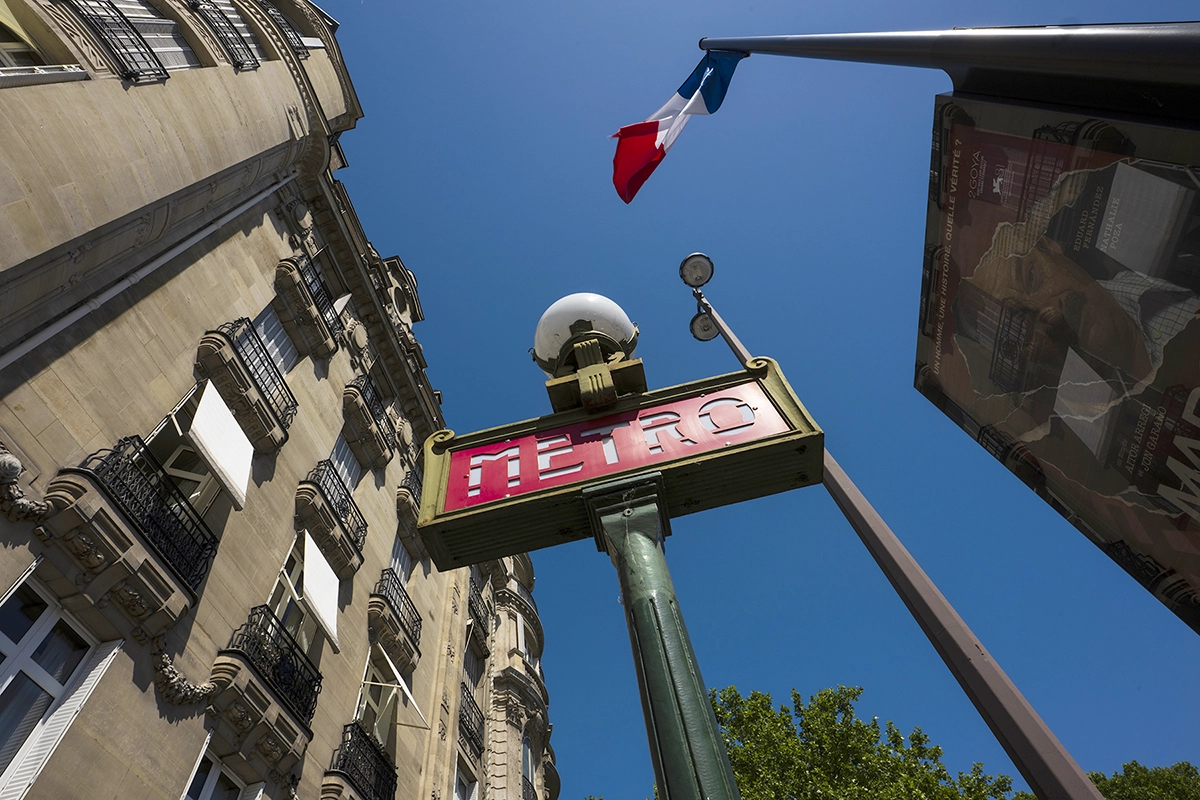 A low-angle view of a red and green "METRO" sign mounted on a tall green pole, against a clear blue sky. A French flag is visible flying from a pole above the sign and in the background, there is a multi-story building with balconies.