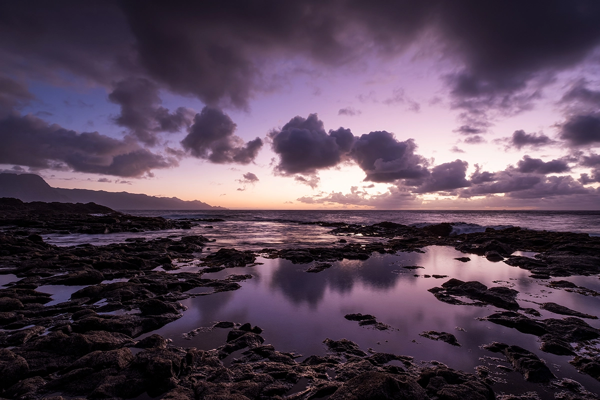 Dramático pôr do sol púrpura sobre formação rochosa de lava e água, fotografado com lente grande angular Tamron para câmeras Fujifilm X.