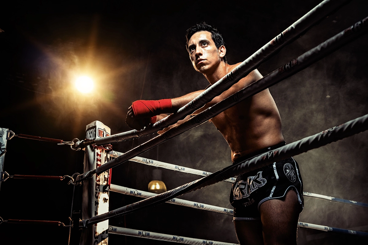 Un luchador de Muay Thai con vendas rojas en las manos se apoya en las cuerdas de un ring de boxeo poco iluminado, mirando intensamente hacia delante. Captado con el Tamron 17-70 mm F2,8 en una cámara Fujifilm con montura X, muestra profundidad, nitidez y dramatismo.
