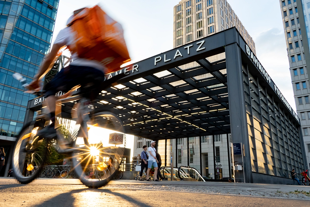 Um ciclista com uma bolsa de entregas laranja passa pela estação Potsdamer Platz, em Berlim, ao pôr do sol, capturando o movimento e a energia da fotografia urbana de rua.