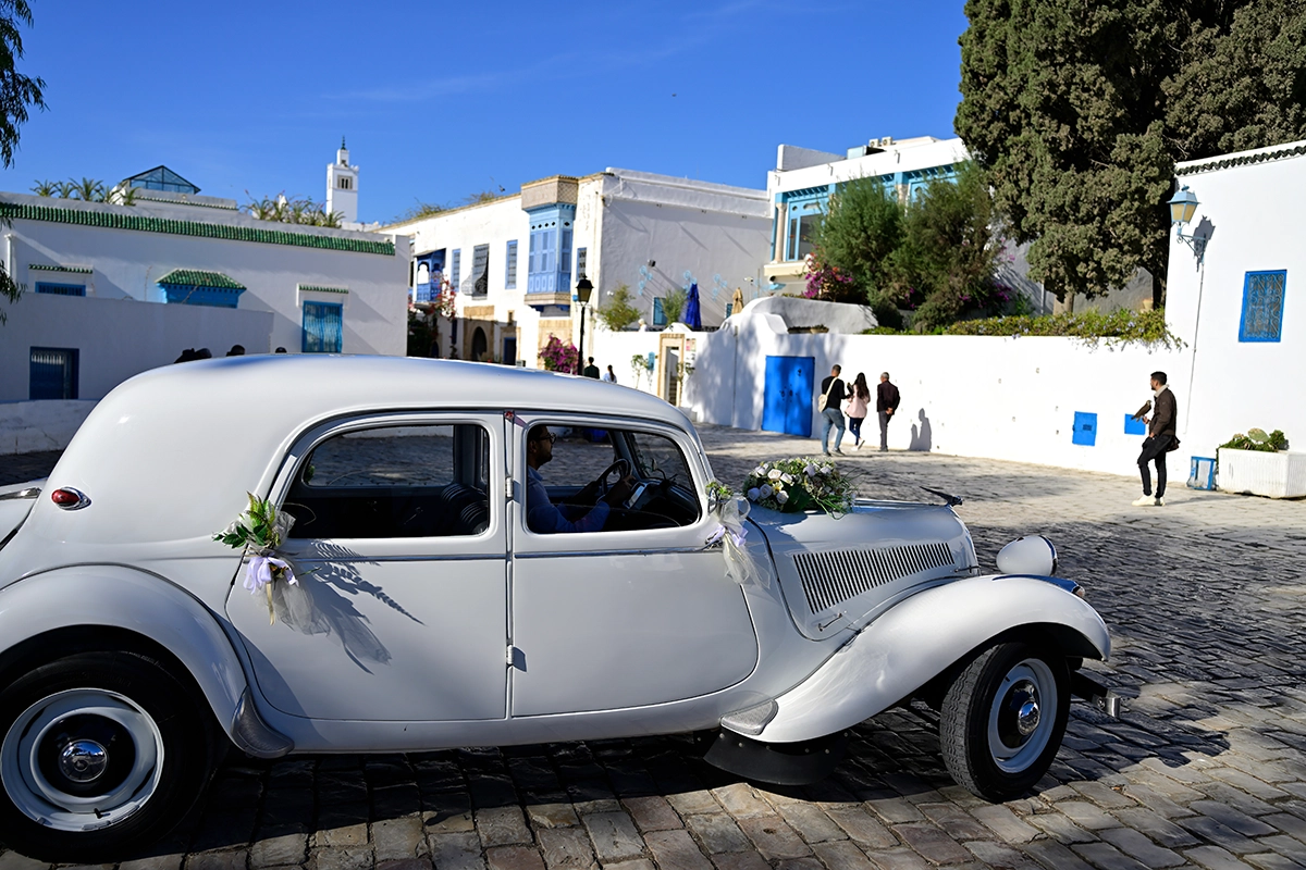 Um carro de casamento branco vintage decorado com flores passa por uma rua mediterrânea iluminada, um exemplo de fotografia de rua que captura a cultura e a celebração.