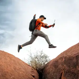 Photo from a low angle of Seth Macey jumping over rocks with a backpack of gear and his camera and Tamron lens in hand.