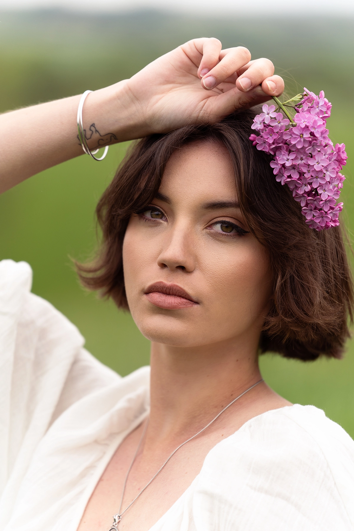 Close-up outdoor portrait of a woman holding lilac flowers above her head, photographed with the Tamron 70-180mm F2.8 G2 lens showing smooth background bokeh.