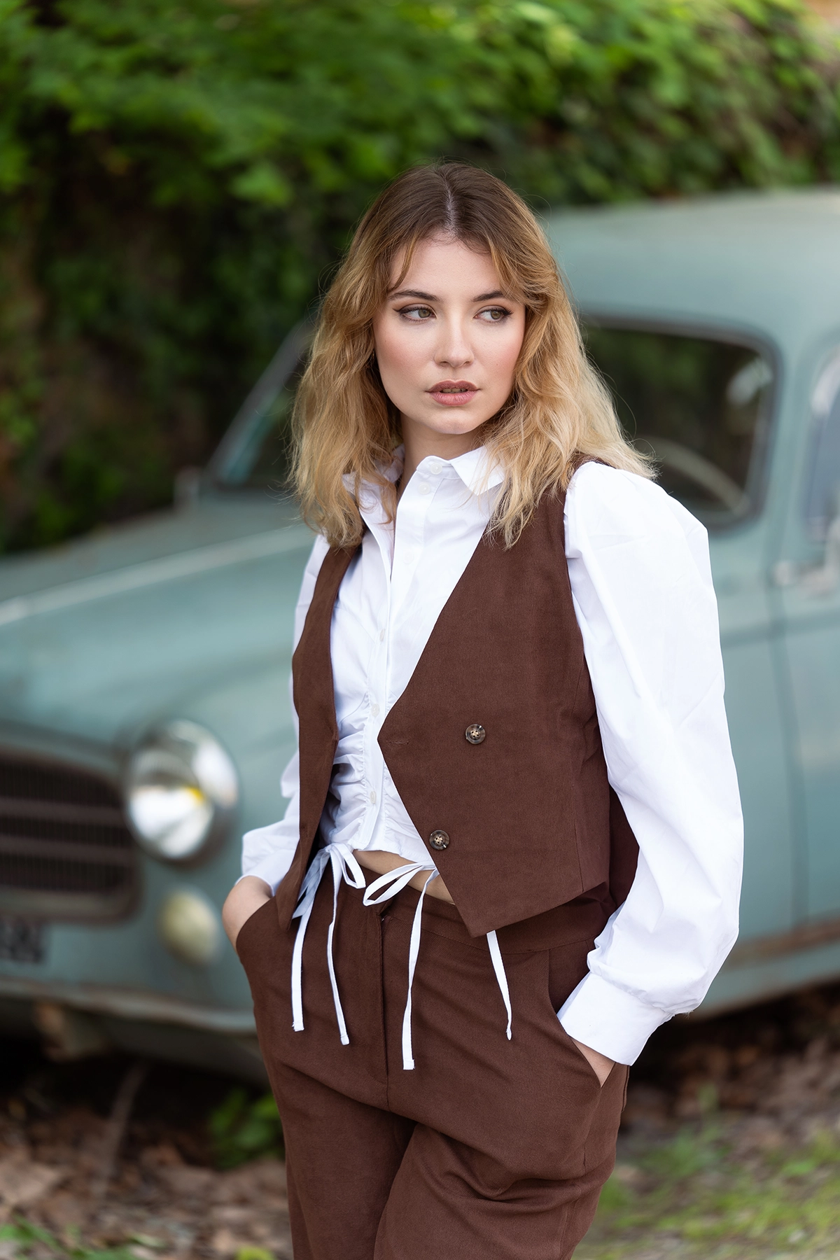 Outdoor portrait of a woman standing in front of an old blue car, photographed with the Tamron 70-180mm F2.8 G2 lens showing shallow depth of field.