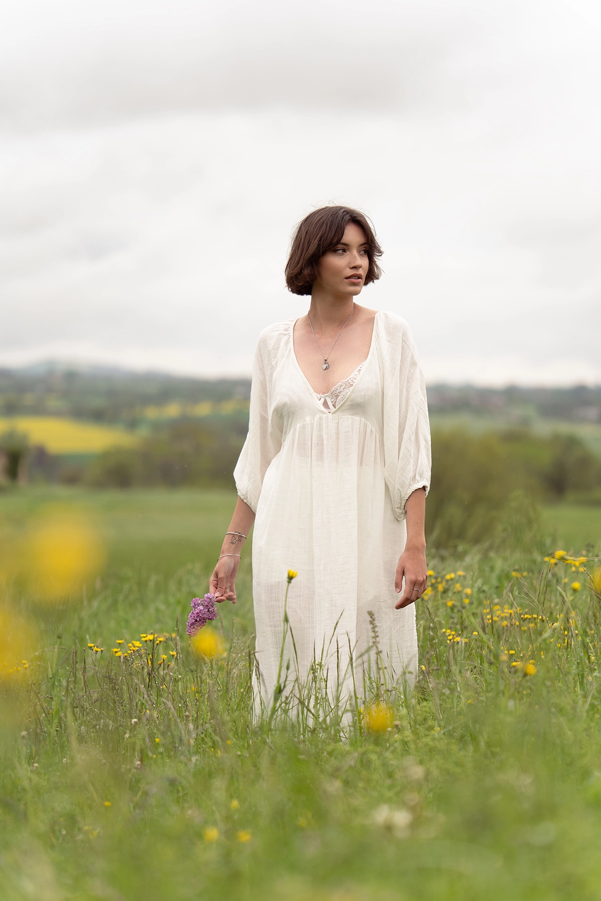Woman standing in a field holding lilac flowers, photographed with the Tamron 70-180mm F2.8 G2 lens for a soft, natural portrait.