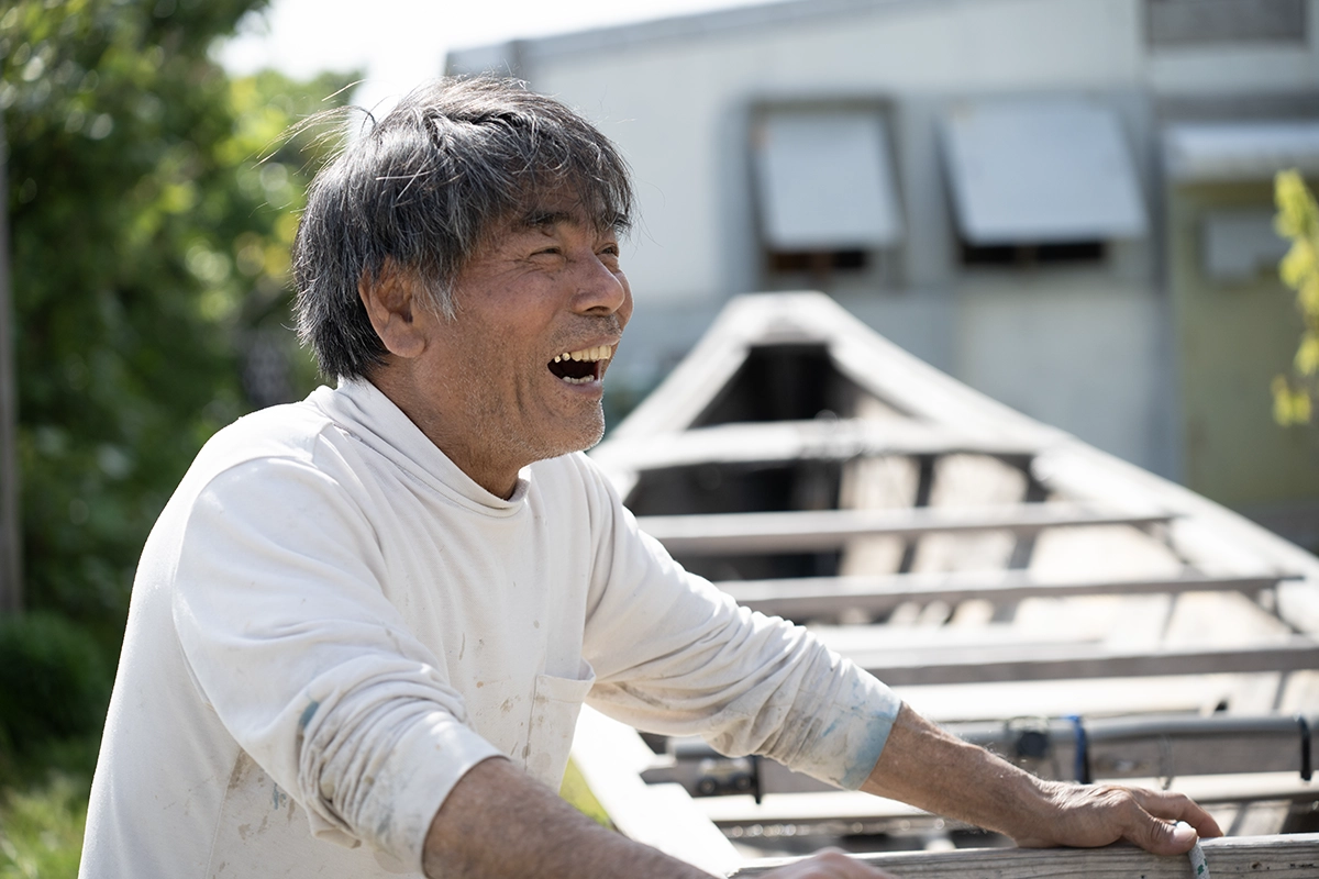 Retrato de um construtor de barcos SABANI de Okinawa rindo ao ar livre ao lado de uma estrutura de casco de madeira, capturado com a Tamron 28-75mm F2.8 G2 para um documentário de narrativa visual.