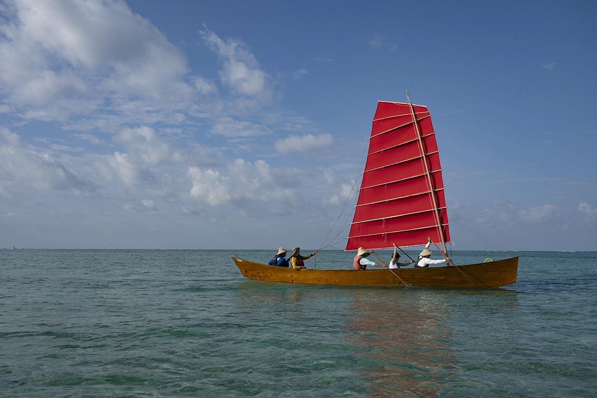 Barco SABANI tradicional de Okinawa com uma vela vermelha brilhante deslizando em águas azuis claras, fotografado com a Tamron 28-75mm F2.8 G2 para um documentário de narrativa visual cultural.