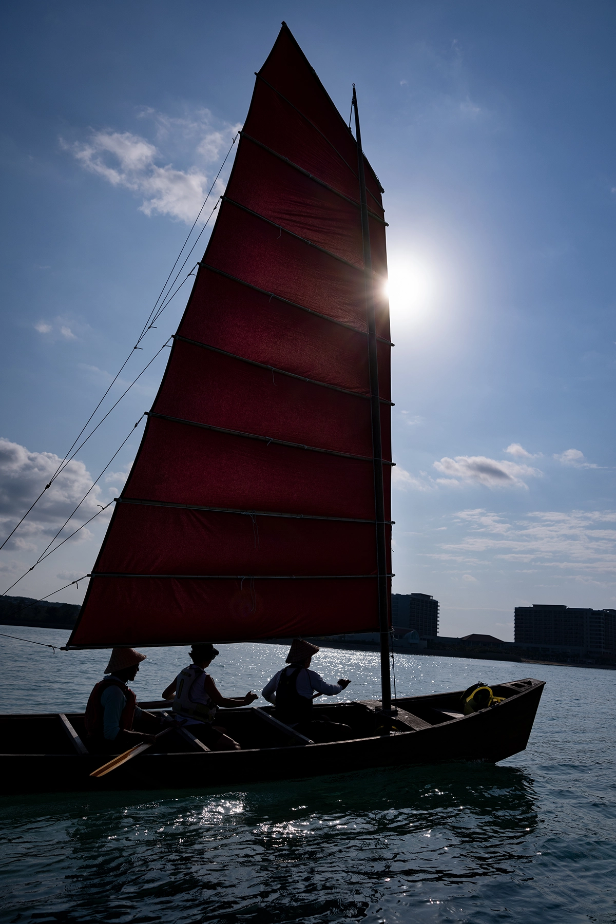A tripulação do barco SABANI em silhueta remando sob uma vela vermelha alta, enquanto a luz do sol incide por trás do mastro, fotografada na costa de Okinawa para um documentário de narrativa visual.